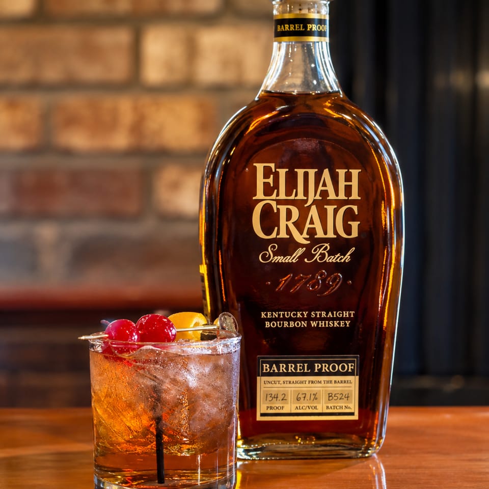Elijah Craig Barrel Proof bourbon bottle beside an iced bourbon cocktail garnished with cherries and an orange slice on a wooden bar.